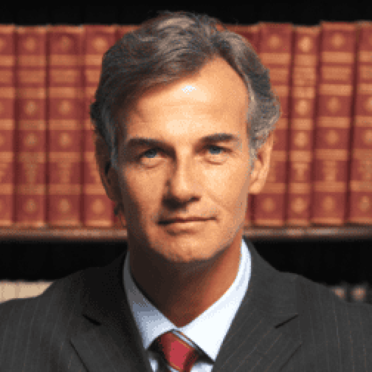 Man in a suit with gray hair stands in front of a row of books on a shelf.